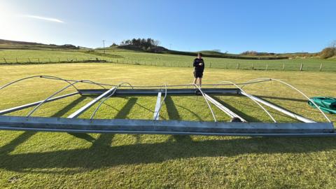 A grey metal frame is lying on a green field. It looks like it has been bent out of shape. There is a blue sky in the background and a person in a black top and shorts is stood behind the frame.
