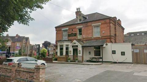 Streetview image of the Sportsman pub, a substantial brick Victorian building standing along in an urban setting with parking area in front