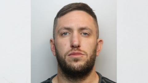 Custody image of Aaron Winzar. He is staring directly in the camera. He has facial hair.