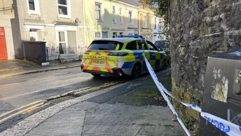 A Devon and Cornwall Police car parked at the side of a road. There is police tape from its door handle to an electricity box. The cordon is across the pavement.