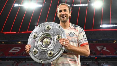 Harry Kane is holding the German Bundesliga championship title bowl. He is wearing a white Bayern Munich jersey.