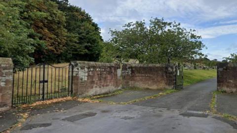 A sandstone wall entrance to Grange Cemetery with two black metal gates