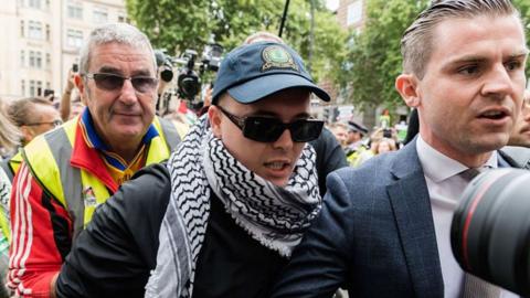 Liam Óg Ó hAnnaidh (wearing a blue keffiyeh , dark blue cap and sunglasses arrives at court. To his right is his solicitor and to his left is a man in  a hi-iz hacket