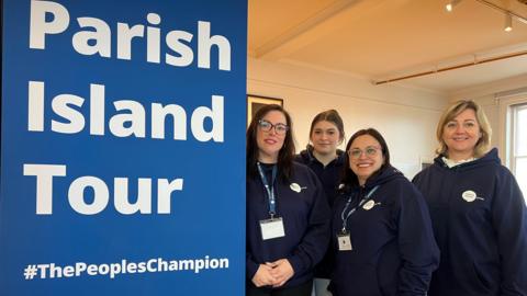 Four women from Citizen's Advice Jersey are smiling at the camera. They are wearing navy branded hoodies and lanyards and are standing next to a big pop-up bright blue banner with white lettering that reads Parish Island Tour #ThePeoplesChampion