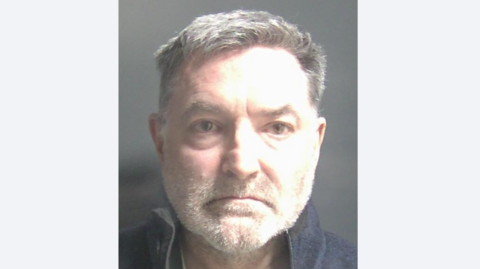 A police mugshot of Derek Johnson wearing a dark grey jumper and standing against a dark grey background. He is a 59-year-old man with grey hair and a short, almost white, beard. He is looking directly at the camera with a blank expression. His brow is furrowed and his mouth is turned down at the corners. There are bags under his eyes.