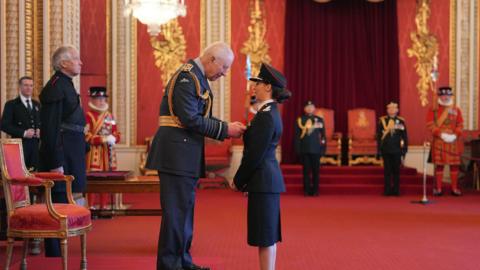 The King presenting Dr Cohen-Hatton with her service medal. They are in a red room and both are wearing black suits. There are people in the background. 