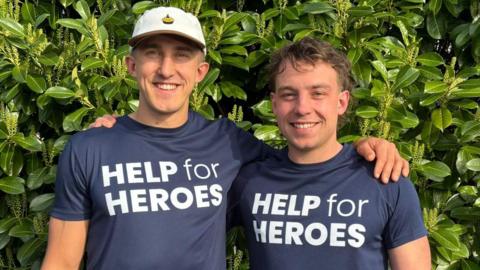 Two young men standing in front of some foliage, their arms around each other. The one on the left is taller and wearing a white baseball cap while the other has short brown hair. Both are wearing dark-blue t-shirts with "HELP FOR HEROES" written on them in white lettering.