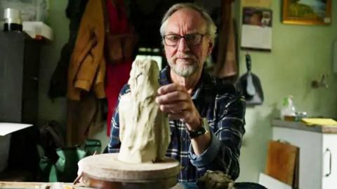 A sculptor with his hand on a sculpture of a person with long hair.