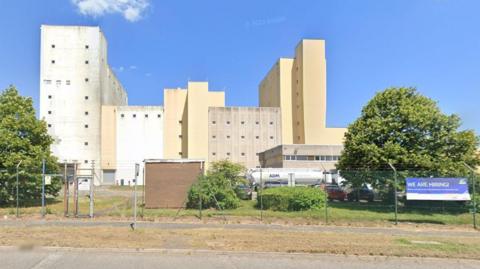 Large concrete building with several storeys. There are towers on either side with a shorter section in the middle. There is a large number of small square windows on the building.  The letters ADM are visible on a smaller white building in the foreground. There are trees either side of the building.