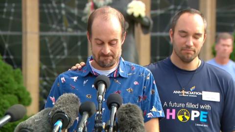 A man in a blue printed button up shirt speaks into several microphones with his head bowed down.