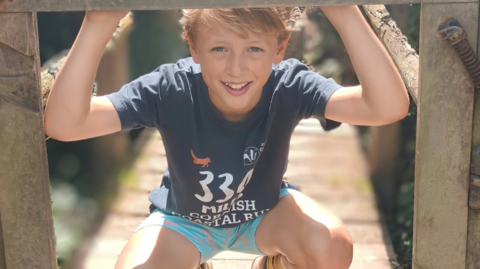 Freddie in a summer t-shirt with 330 miles Cornish Coastal Run logo, and turquoise shorts, bending down at a wooden bridge and smiling to the camera