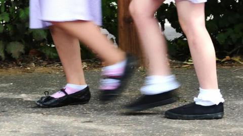 A generic image showing the legs of two primary school girls wearing ankle socks and black shoes on a path. They are out of focus as if the girls are skipping or running