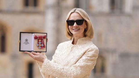 Carey Mulligan holds up a CBE medal in a box while posing for a photo in the grounds of Windsor Castle. She has a blonde bob, and is wearing sunglasses and a cream embroidered jacket.
