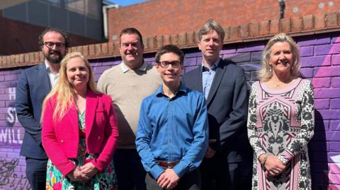 Six smartly-dressed candidates for Huntingdonshire District Council elections pose for a photograph. Two woman and one man are on the front row, and three men are on the back row. They are stood in front of a purple graffiti-d wall.