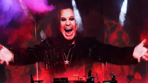 An huge picture of Ozzy Osbourne, with his mouth wide open in his trademark snarl, wearing black and his arms outstretched. The image is bathed in smoke and red and purple lights above musicians performing on stage.