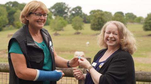Dr Tania Gilbert smiling while receiving the award by a representative of The British and Irish Association of Zoos and Aquariums. The other woman is also smiling. They are outside, in what it looks like part of the zoo. They are both looking at the camera.