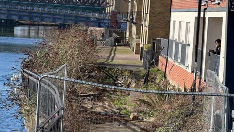 A fenced off footpath with a sinkhole in it- placed between the River Nene and a block of flats on the other side of it.