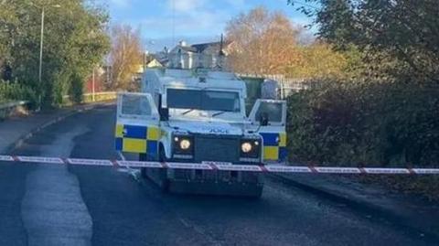 A PSNI landrover on an empty road, which has trees on either side. Its two side doors are open and it sits behind some police tape.