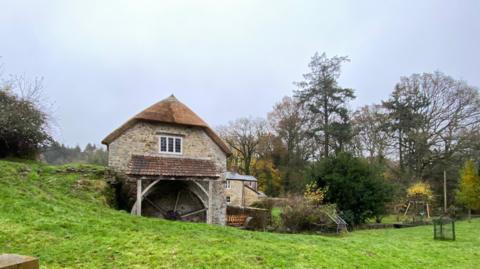 A stone mill is the focus of the picture with the wooden wheel visible underneath a tiled roof. There are other stone buildings behind it with gardens. One has a child's swing in the garden. The grass in front of the mill is a very vivid green colour while the sky is a very pale blue.