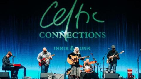 A band performing onstage, with the backdrop saying Celtic Connections in large letters. Emmylou Harris is centre stage - a woman in a black dress with blond/white hair, playing an acoustic guitar. Other musicians playing guitars, keyboards and drums are also on stage.  