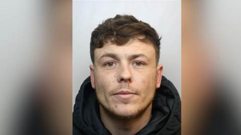 A police mugshot of Lewis Hobbs. He is wearing a black hooded coat and has brown wavy hair and stubble.