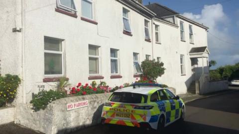 A white two-storey building with several windows and a small garden area in front. A police car is parked outside.