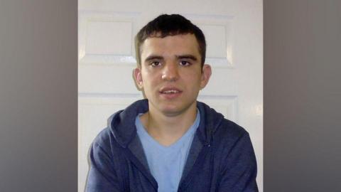 Declan Morrison with short black hair pictured in front of a white interior door. He is wearing a pale blue T-shirt and navy hoodie.