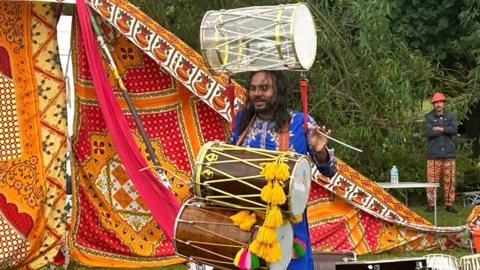 A colourful scene with a backdrop of colourful cloth and a dancer with several drums in the foreground. 