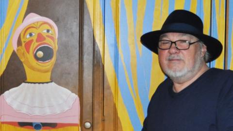 Pete Sinfield wearing a fedora-style hat standing in front of a wardrobe which has a picture of a fairground clown game painted on it