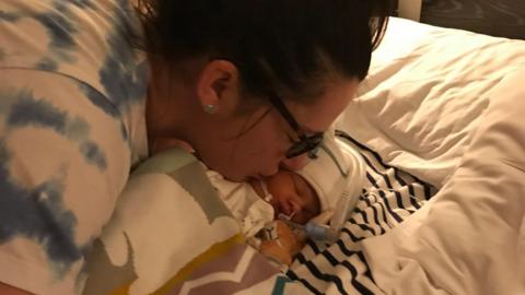 Sarah Robinson, who has dark brown hair pinned up and brown glasses, kisses her baby daughter Ida Lock on the cheek. The baby girl is wearing a white hat and is attached to tubes and wires.