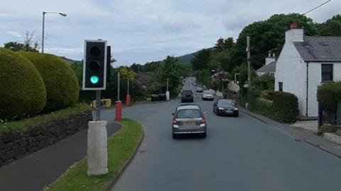 The traffic lights at the junction in Marown. The light is green and there are cars on the road, with houses on the right and green shaped hedges on the left.