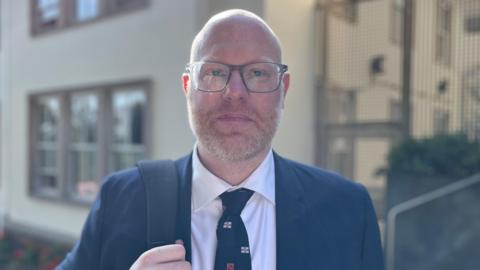 A bald man in a suit looking directly to the camera. He is wearing a dark blue suit with a white shirt and a tie with Guernsey flags on.