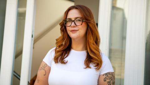 Jess King, a woman with long ginger hair and glasses, stands in front of a doorway. She is wearing a short-sleeved white top and has visible black tattoos at the top of her arms. 