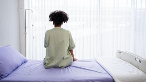 A person in a pale green hospital gown sits on a hsopital bed, made up with a lilac bedspread, pillow case and white blanket. They are facing away from the camera towards the window, with a gurney left of them. 