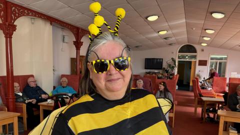 A woman in a fancy dress bee outfit and black and yellow sunglasses smiles at the camera , behind her are a group of elderly people sitting in chairs at a day centre 