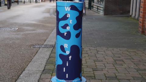 A blue cylinder shaped water filling station. It says water refill vertically in white letters