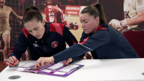 Katie Bradley and Jodie Hutton dressed in Charlton Athletic tracksuits, play with the stickers on a table.