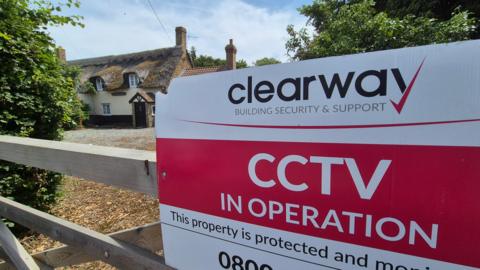A white and red sign reads "Clearway Building Security and Support - CCTV in operation". The sign is attached to a wooden gate, with a thatched cottage and trees in the background