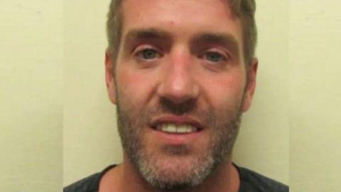 A police mugshot of a smirking Jamie Rothwell, who has dark stubble tinged with grey and light brown.