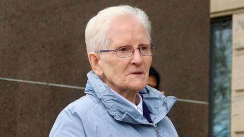Head and shoulders shot of Marie O'Gorman, who has short grey hair swept back, outside Glasgow Sheriff Court. She is wearing a light blue jacket with a large collar and has dark-rimmed glasses. She is looking ahead, to the right of the photographer.