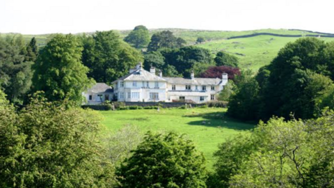 A view of High Borrans, Windermere, an outdoor education centre. The building is large and white with a slate roof and is surrounded by green fields and large trees.