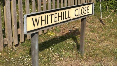 A road sign saying Whitehill Close, on wooden posts with grass and small white flowers around its base