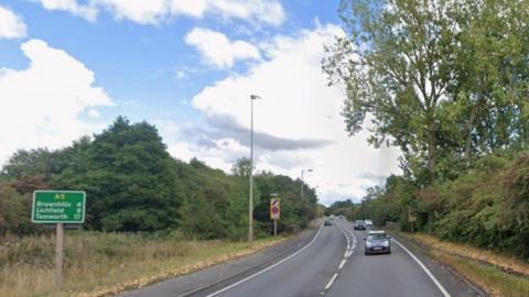A road with vehicles travelling in both directions. There are street lights and green road signs at the side of the road as well as a pavement running for some of its length.