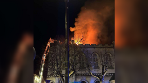 Flames and smoke seen against the night sky and firefighters alongside the burning building on an aerial platform, spraying water on the flames.