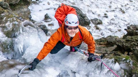 A climber wearing an orange jacket, helmet, and gloves ascends a frozen waterfall using two ice axes. A pink rope is clipped into their harness, and a red backpack rests on their back. The icy rock face is steep and rugged, with patches of exposed stone.