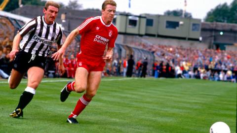 Steve Nicol with all-red football kit is chasing a ball pursued by a Newcastle player in black and white stripes with black shorts and black socks with white tops.