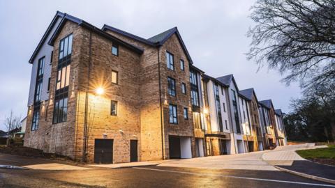 A large brown-brick apartment complex, with outside lights on in the middle of the front of the building. There are pitched roofs along the complex with some trees to the right.