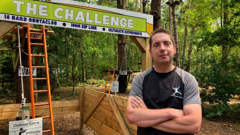 A man in a black and grey T-shirt looks at the camera with his arms crossed. He is standing in front of lots of trees. To his right there is a ladder propped up against a tree and a sign above reads "The Challenge"