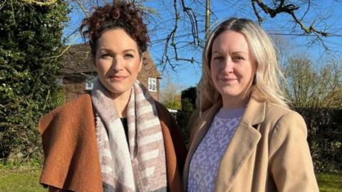 Amira Stallion and Amber Bradly stand side by side with a hedge behind them. Amira is wearing an orange coat and patterned scarf. Her brown curly hair is tied in a ponytail. Amber is wearing a cream coat and has blonde mid length hair. 