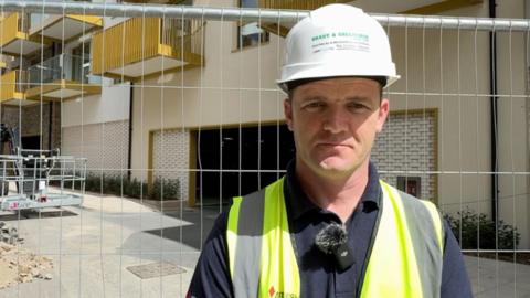 Martin Burns, a man, wearing a white hard hat which says 'Brady & Gallagher' in green letters on the front. He is also wearing a yellow / green high-visibility jacket, over a dark blue polo shirt. Behind him is a metal fence and a building site, featuring balconies with mustard yellow railings.